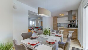 Une cuisine moderne avec des meubles blancs, un coin repas avec une table blanche dressée pour quatre personnes, et un salon avec un canapé et une table basse.