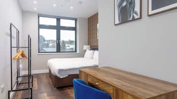 Quarto de hotel com cama, escrivaninha, cadeira, guarda-roupa, janela com vista para a cidade e cabideiro com cabides.