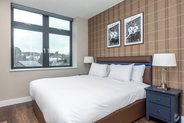 A neatly made bed with white linens, a headboard with a blue accent, and a nightstand with a lamp. Two framed pictures hang on the wall above the bed.