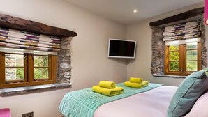 A bedroom with a bed, two windows with stone frames, a television on the wall, and a pink chair. - The Cosy Peacock (Windermere)
