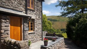 A stone building with a wooden door and windows, a stone pathway leading to a seating area with potted plants, and a view of hills in the background. - The Cosy Peacock (Windermere)