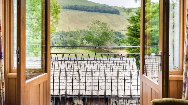 A room with a wooden door leading to a balcony with a metal railing, offering a view of a hilly landscape. - The Cosy Peacock (Windermere)
