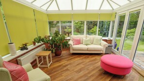 A conservatory with a wooden floor, a beige sofa, a pink ottoman, and a green armchair. - Number 16 (Woodhall Spa)