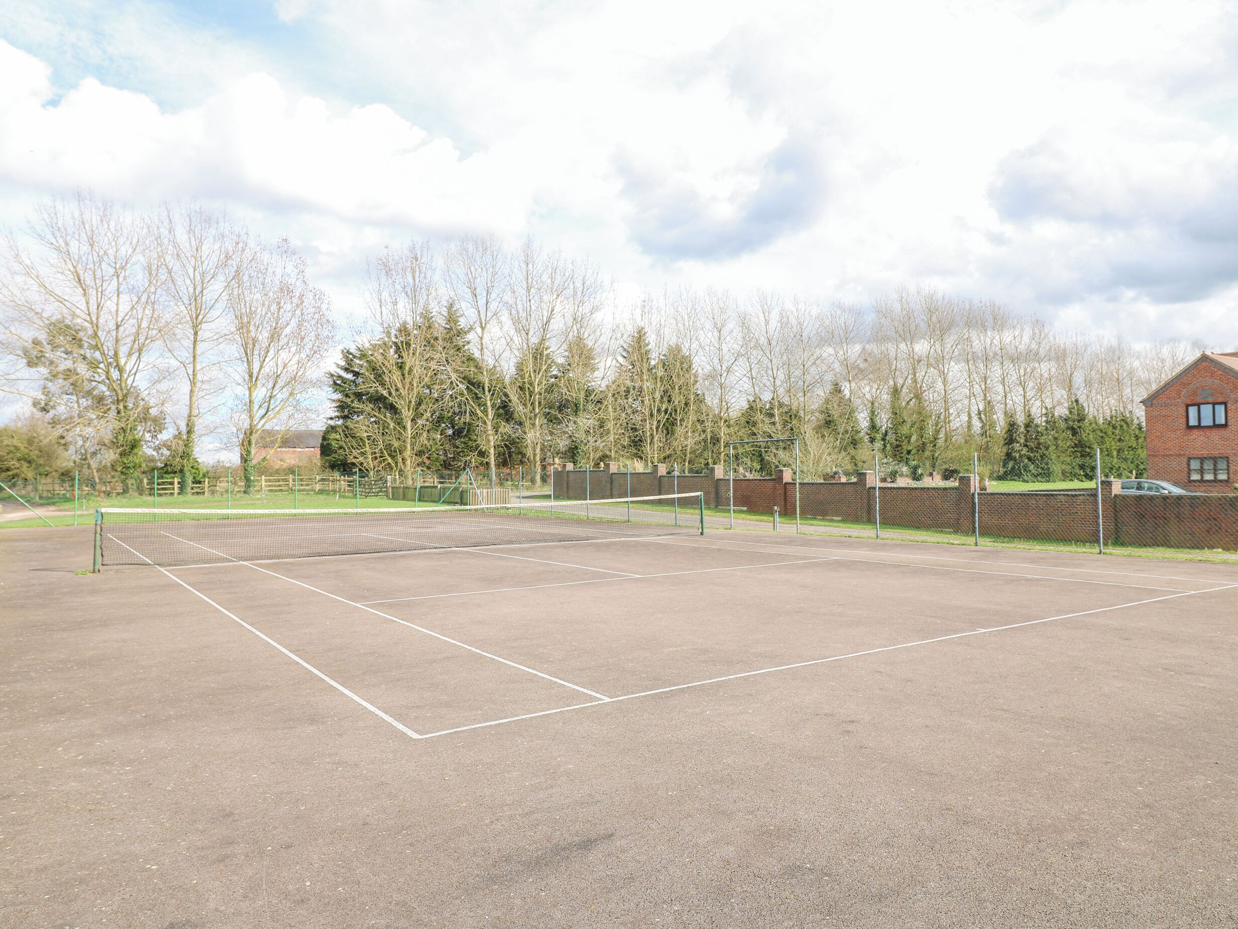 Cancha de tenis exterior con red, rodeada de árboles y un edificio de ladrillo.