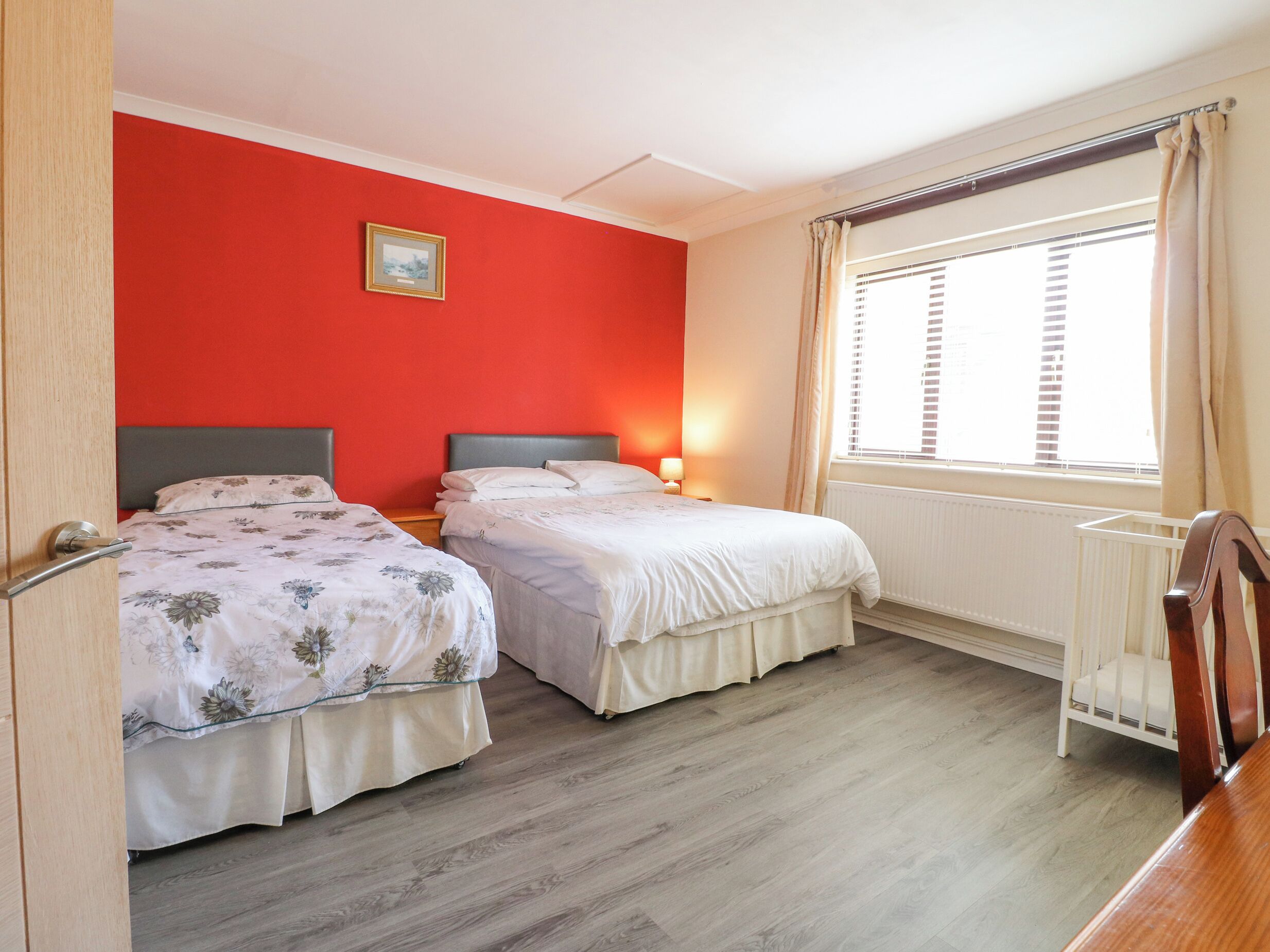 A room with two beds, a wooden floor, a red wall, a window with blinds, and a crib.