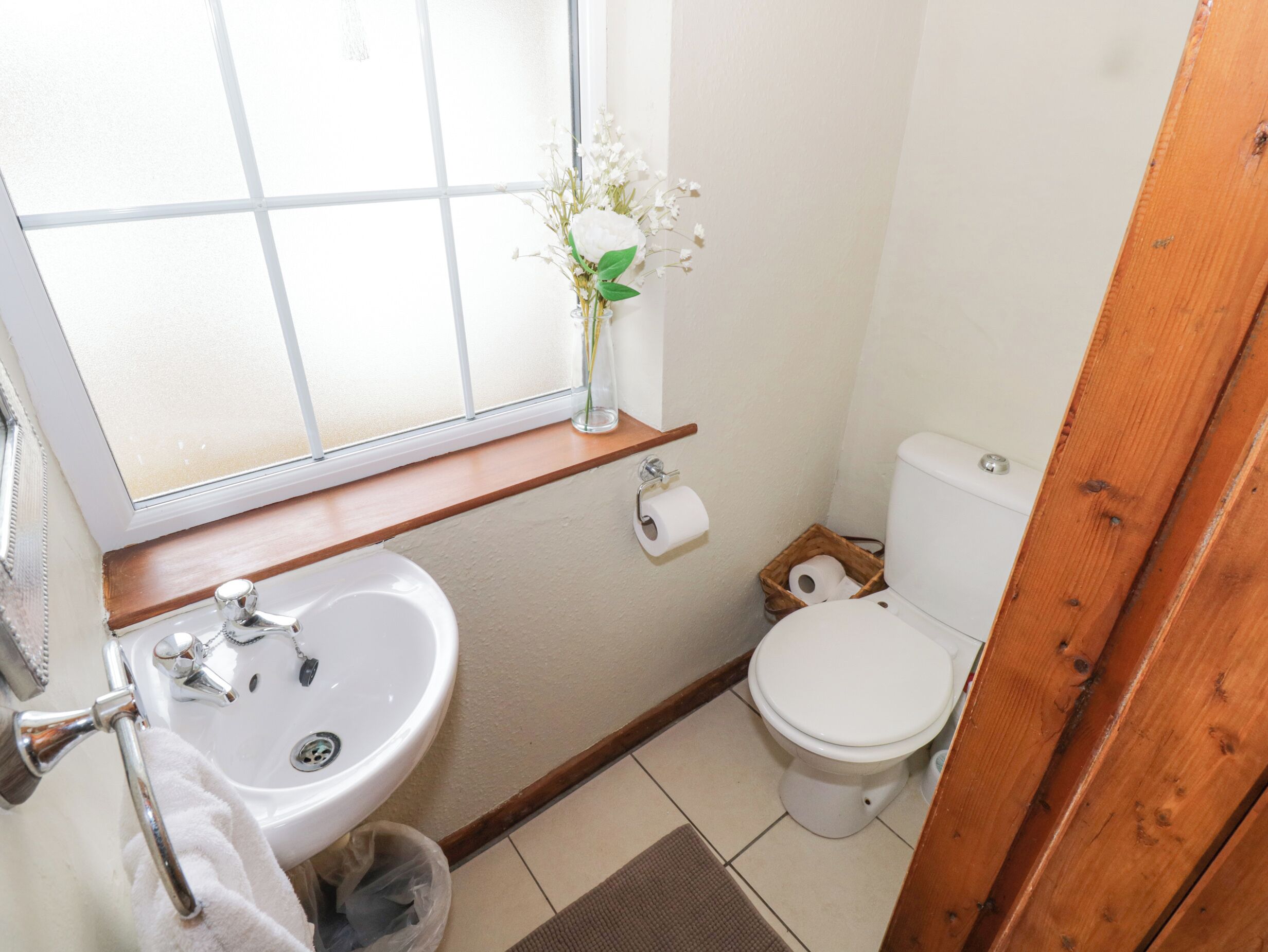 A small bathroom with a window, sink, toilet, and a wooden shelf with a vase of flowers.
