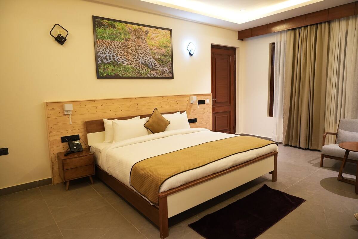 Une chambre d’hôtel avec une tête de lit en bois, un lit recouvert de draps blancs et d’un plaid marron, une table de chevet avec un téléphone, une lampe fixée au mur et un tableau représentant un léopard.