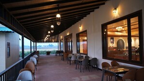 Un área de estar al aire libre con techo, mesas y sillas, una zona de bar con mostrador y taburetes, y grandes ventanales con vistas al paisaje.