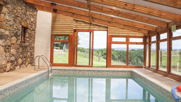 A swimming pool with a stone wall and wooden beams, large windows, and a view of the countryside. - Skiber Coombe (Callington)