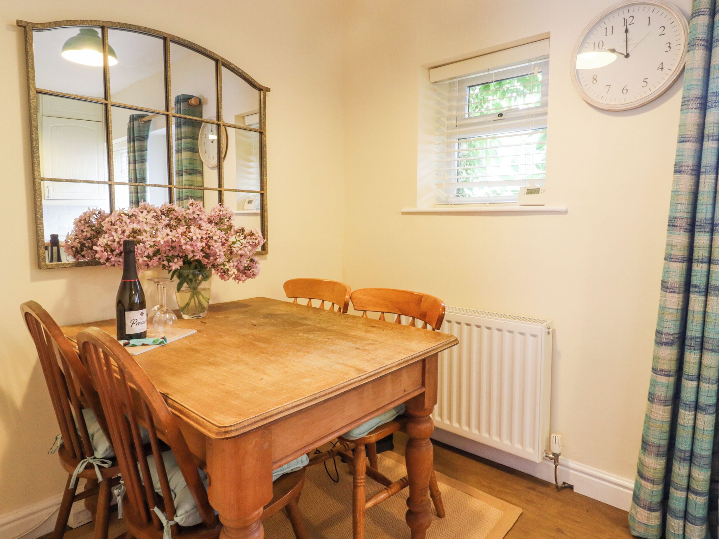 A dining room with a wooden table, chairs, a vase of flowers, a wine bottle, and a clock on the wall.