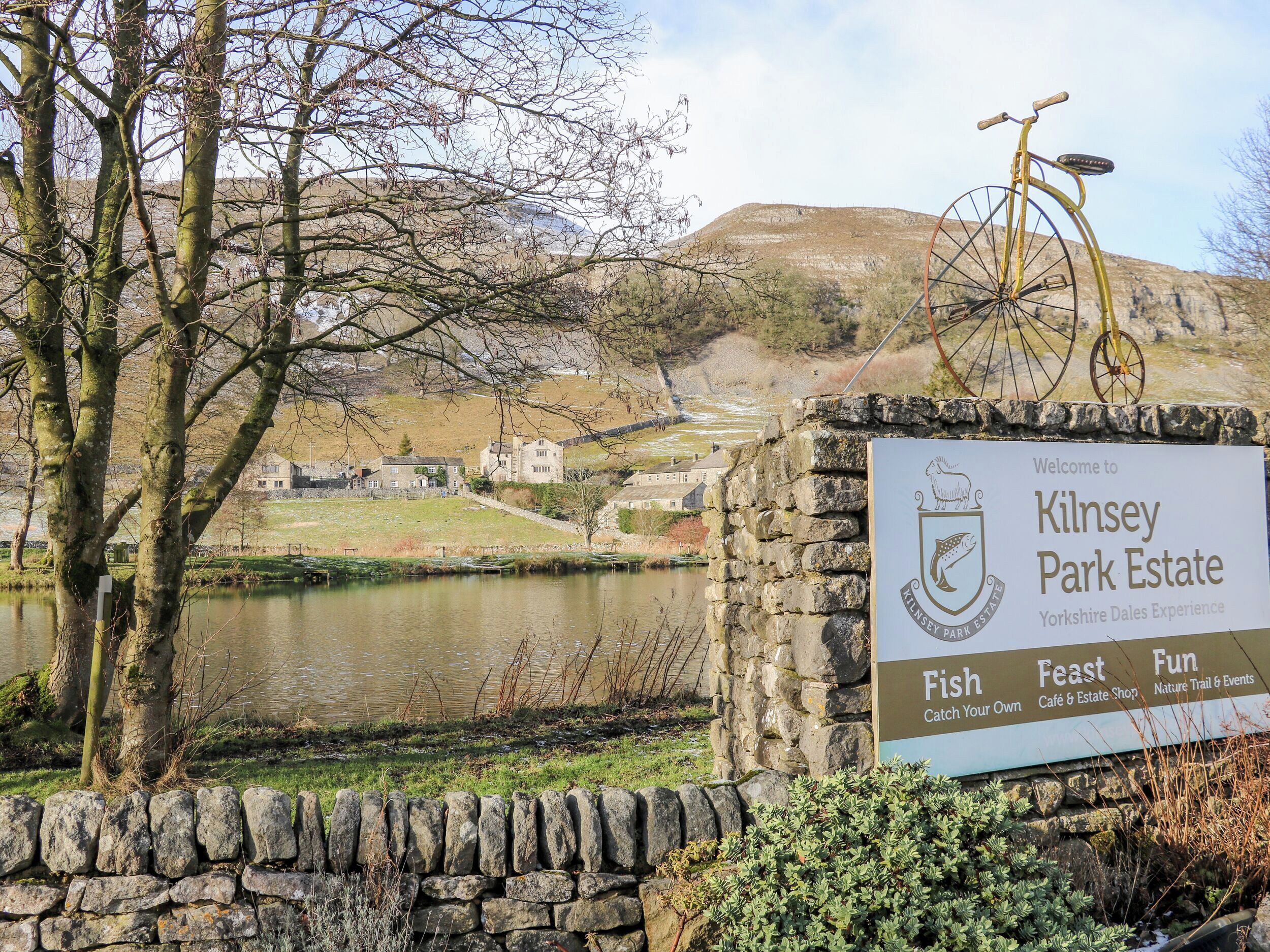 Cartel de Kilnsey Park Estate con vista a un lago, un muro de piedra y una bicicleta.