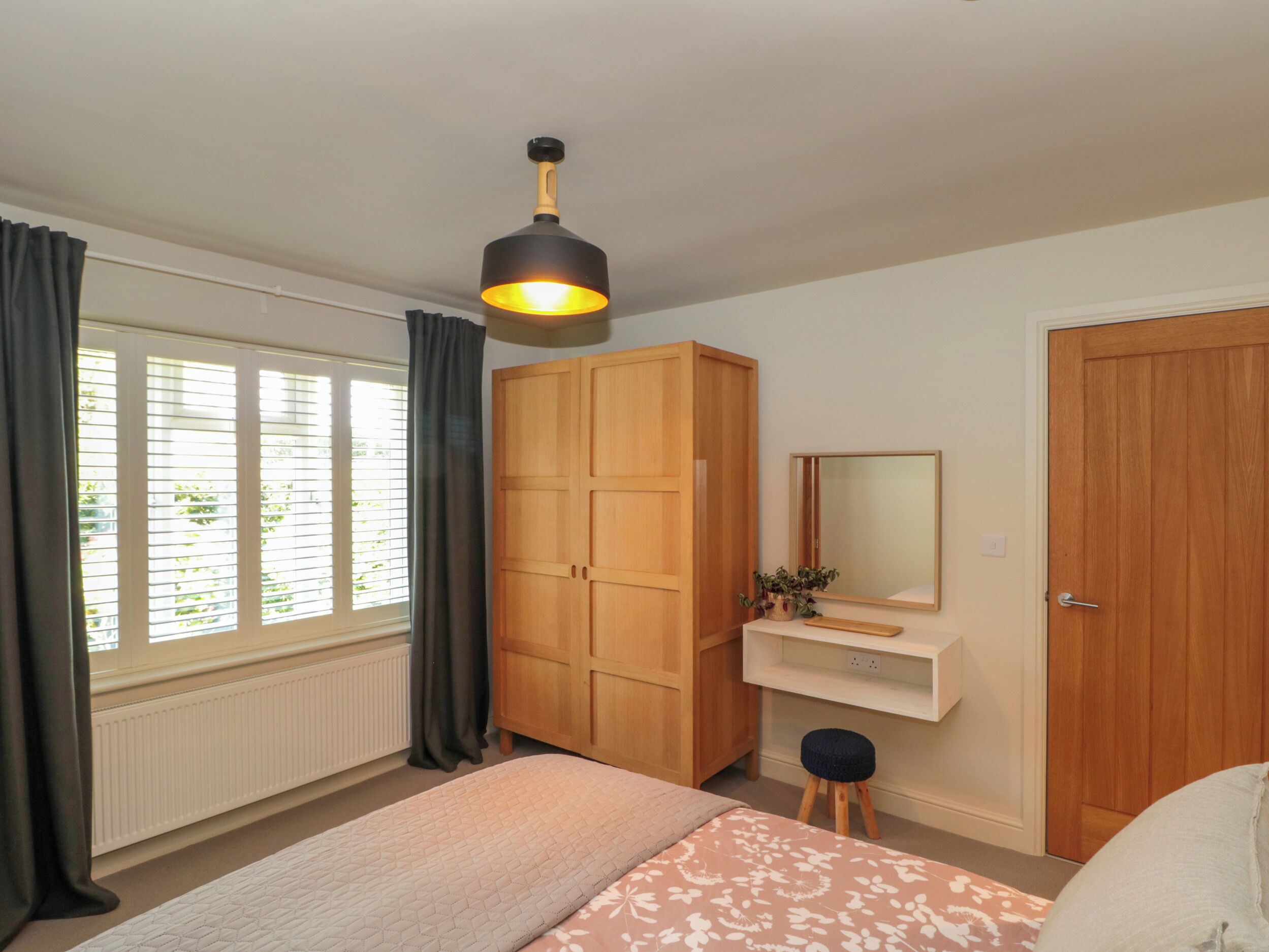 A bedroom with a bed, a wooden wardrobe, a small stool, a mirror, and a hanging light fixture.