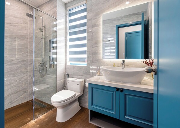 A modern bathroom with a glass-enclosed shower, a white toilet, a large sink with a blue cabinet, and a mirror above the sink.