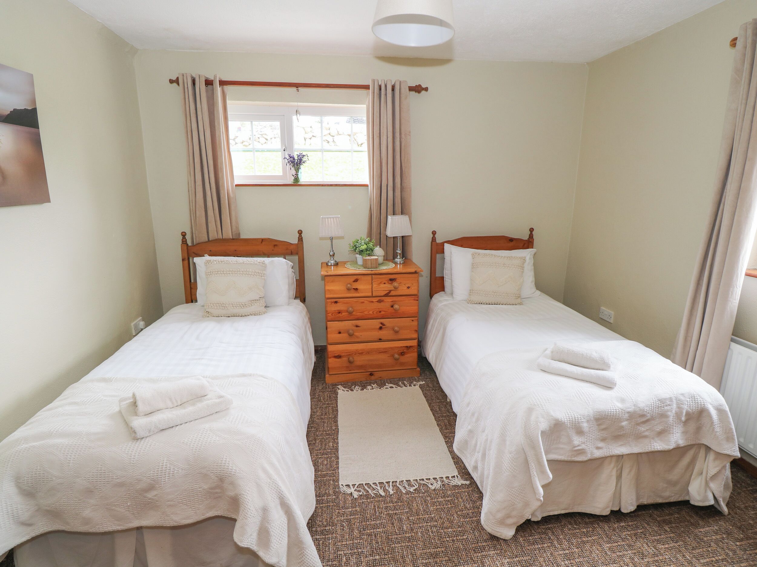 Een slaapkamer met twee eenpersoonsbedden, een houten dressoir, een raam met gordijnen en een klein tafeltje met een lamp en een plant.