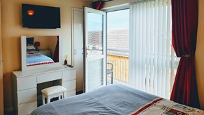 Une chambre à coucher comprenant un lit, une commode, un miroir, une télévision et un balcon avec vue sur les immeubles.
