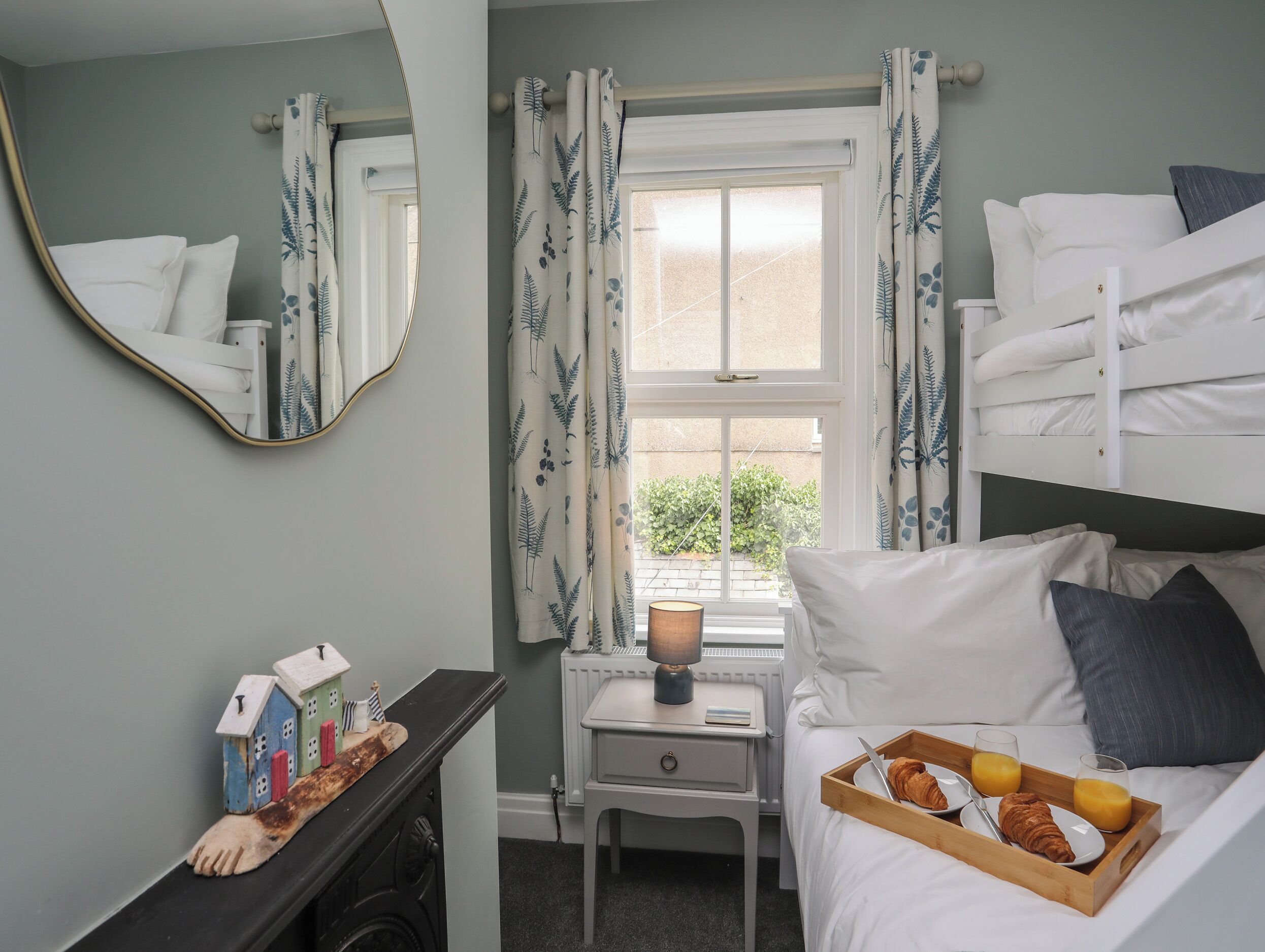 A bedroom with a bunk bed, a bedside table with a lamp, a small desk with a mirror, and a window with curtains.
