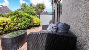 En terrasse med fletmøbler, et bord med glasplade og udsigt til en have med forskellige planter.