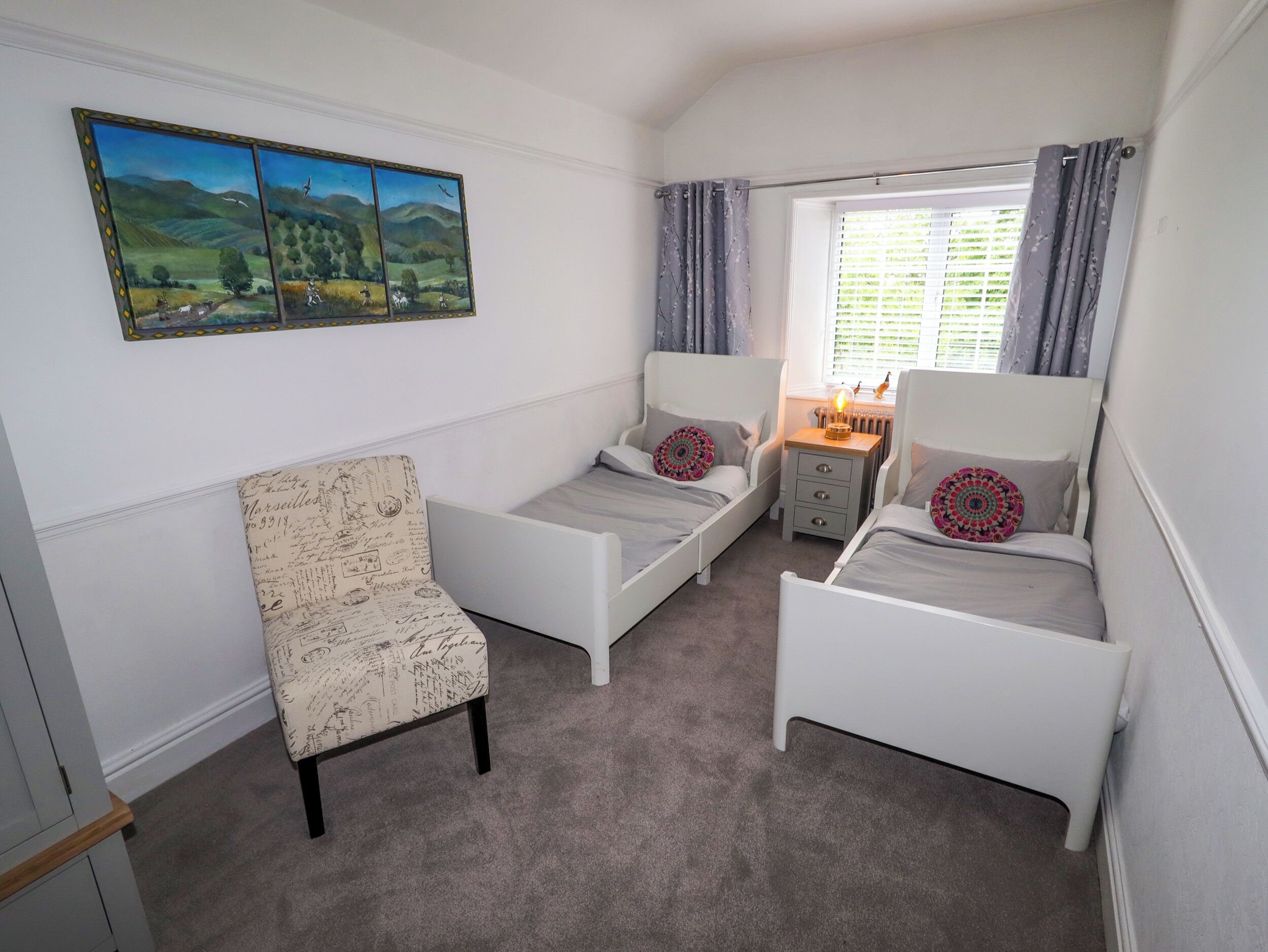 A bedroom with two single beds, a chair, a small bedside table with a lamp, and a window with curtains.