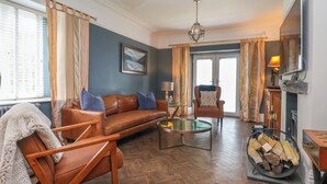 Una sala de estar con piso de madera en patrón de espiga, un sofá de cuero marrón con cojines azules, una mesa de centro de vidrio, un sillón de madera y una chimenea con estufa de leña.