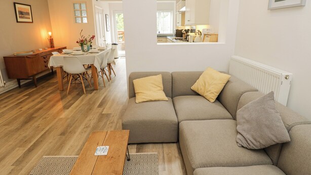 Een moderne woonkamer met een grijze bank, een houten salontafel en een tapijt met dessin. Op de achtergrond is een eetgedeelte met een witte tafel en stoelen te zien.