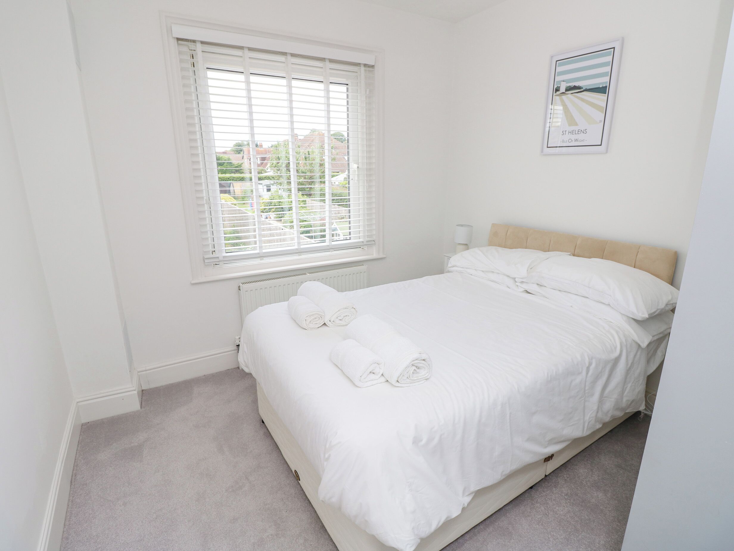Een slaapkamer met een bed, witte beddengoed, een raam met zonwering, een radiator en een ingelijst schilderij aan de muur.