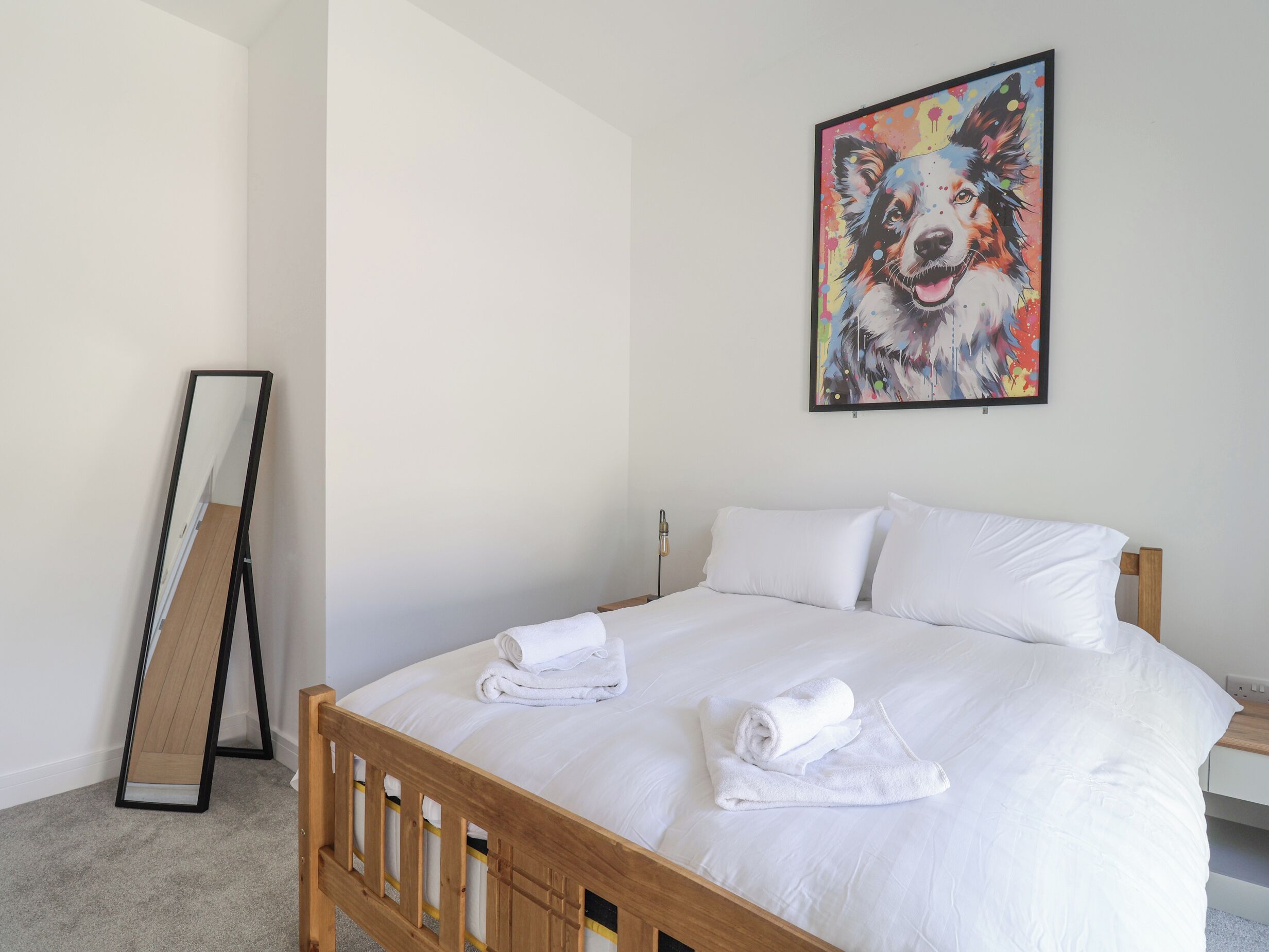 A neatly made bed with white linens and two rolled towels, a colorful dog painting on the wall, and a standing mirror against the wall.