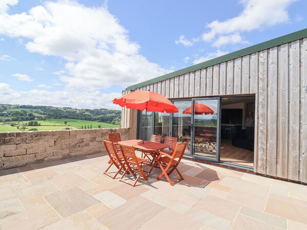 An outdoor patio with a table and chairs, red umbrellas, and a view of the countryside. - Pit Close (Honiton)