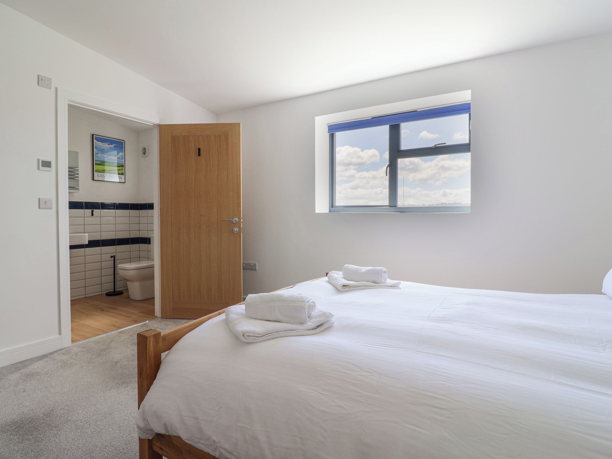 A bedroom with a large bed, a window with a view of the sky, and a bathroom visible through an open door.