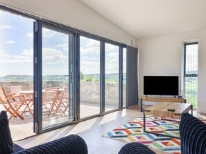 A spacious living area with a large sliding glass door leading to an outdoor terrace, a flat-screen TV, and a colorful rug.
