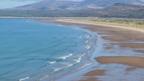 A coastal landscape with a sandy beach, gentle waves, and distant mountains under a partly cloudy sky. - Ael Y Don (Harlech)