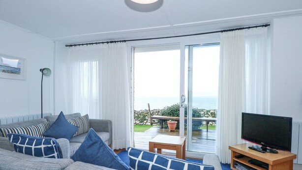 A living room with a grey sofa, blue cushions, a TV on a wooden stand, and a view of the sea through large glass doors. - Ael Y Don (Harlech)