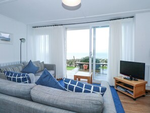 A living room with a grey sofa, blue cushions, a TV on a wooden stand, and a view of the sea through large glass doors. - Ael Y Don (Harlech)