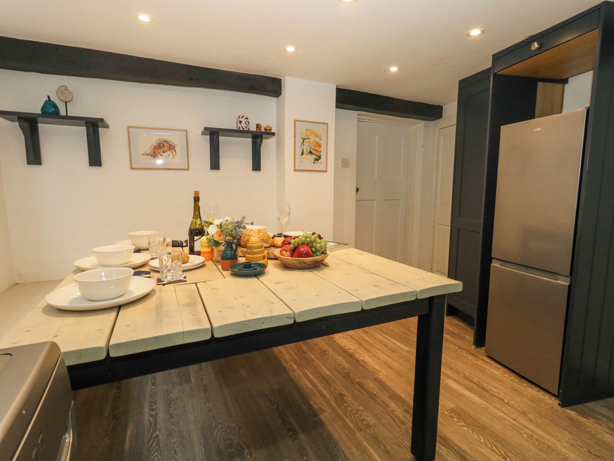 A kitchen with a wooden table set for a meal, a refrigerator, and wall-mounted shelves with decorative items.