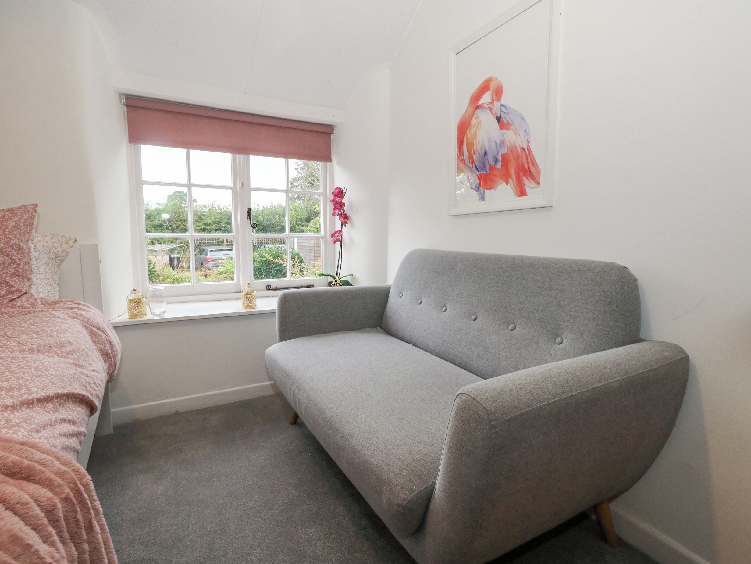 Une chambre avec un canapé gris, un lit rose, une fenêtre donnant sur un paysage verdoyant et un tableau représentant un flamant rose.