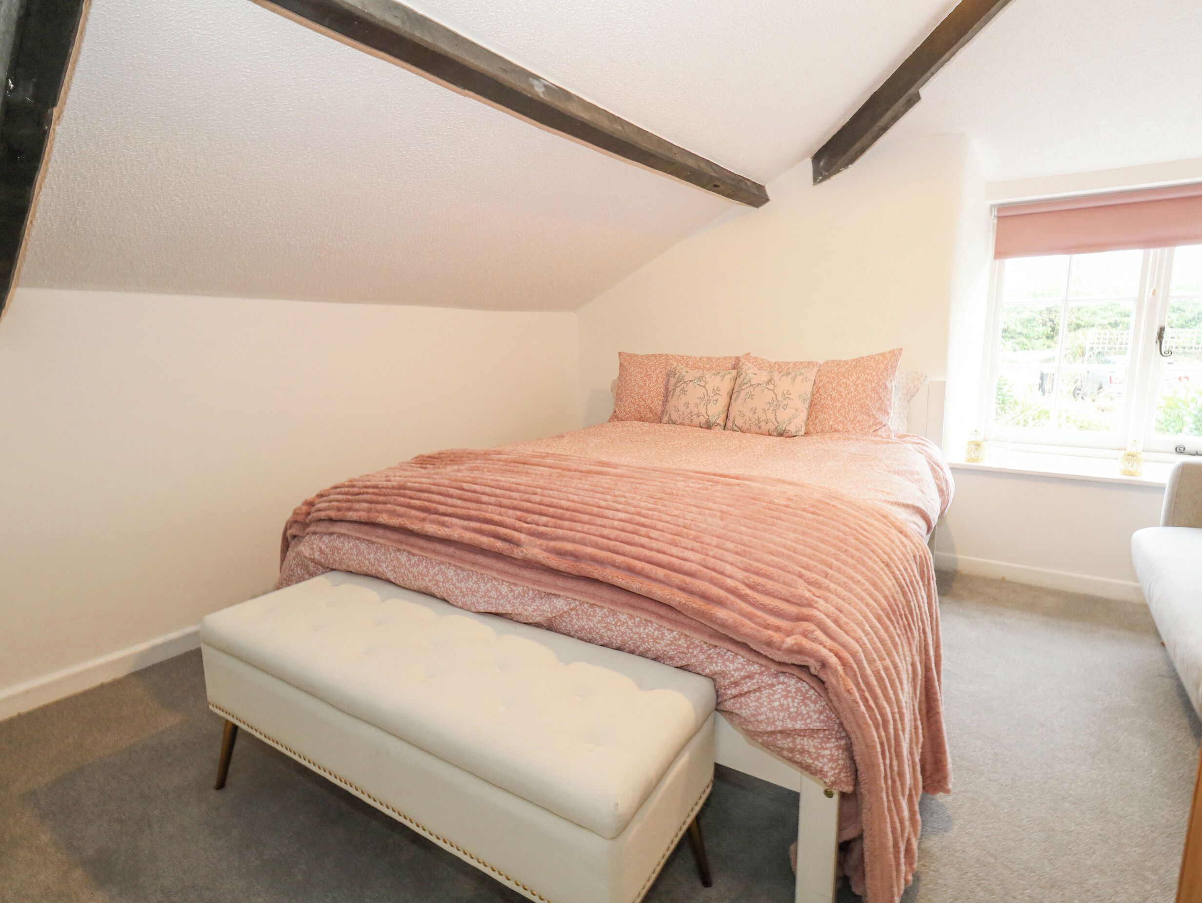 A bedroom with a sloped ceiling, a bed with a pink blanket, a bench, and a window with a view of greenery.