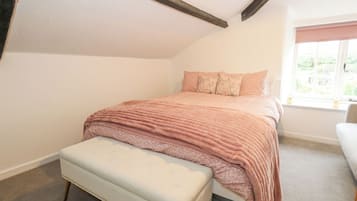 A bedroom with a sloped ceiling, a bed with a pink blanket, a bench, and a window with a view of greenery.