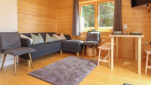Ein gemütliches Blockhaussinnenzimmer mit Sofa, Sesseln, Esstisch, Fernseher und einem Fenster mit Vorhängen.