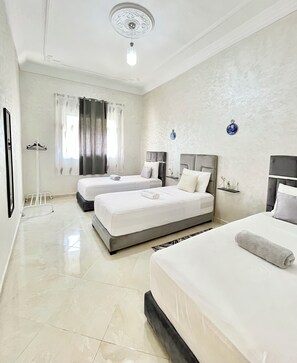 A hotel room with two beds, a hanging light fixture, a window with curtains, and a wall-mounted shelf with a bottle.