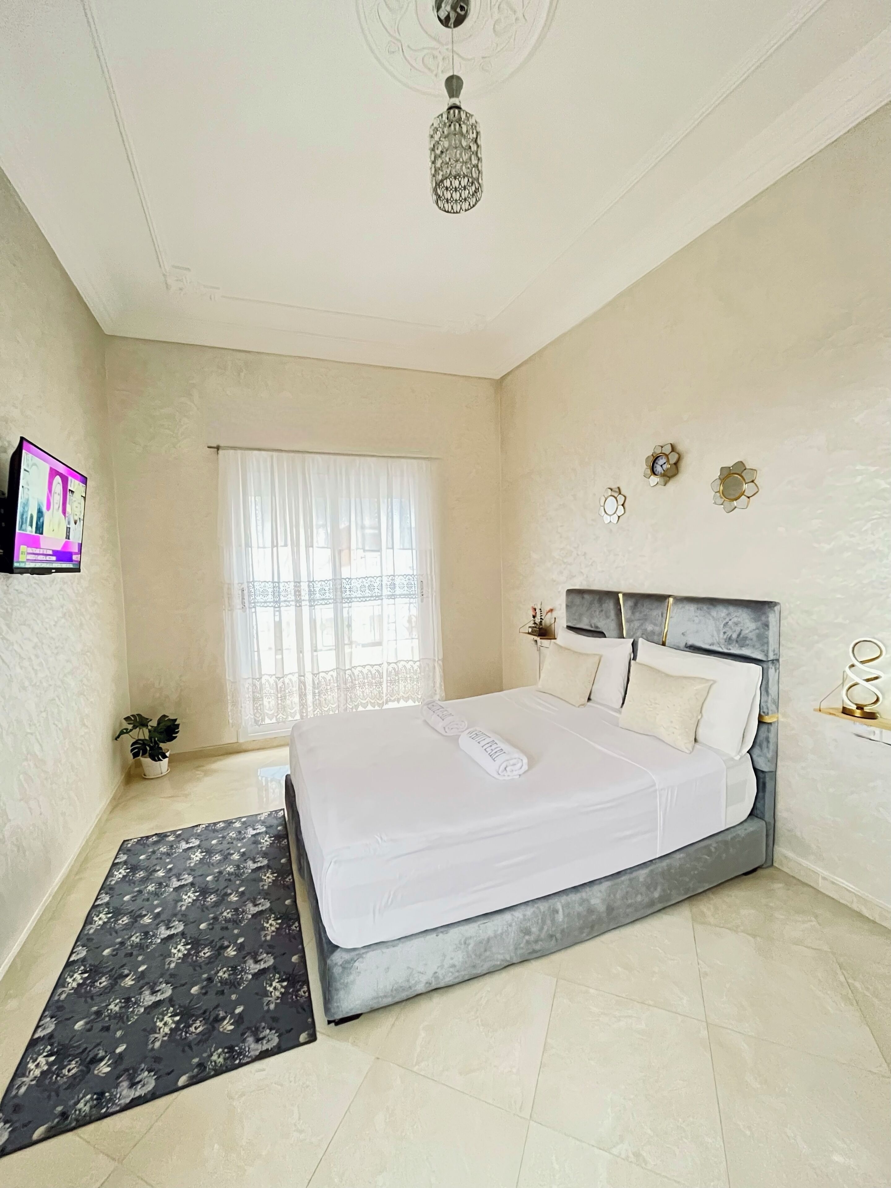 Quarto com cama, televisão, janela com cortinas, planta e lustre.