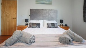 A neatly made bed with a headboard, two bedside tables with lamps, a wooden door, and a painting on the wall. - Apartment 2 (Newquay)