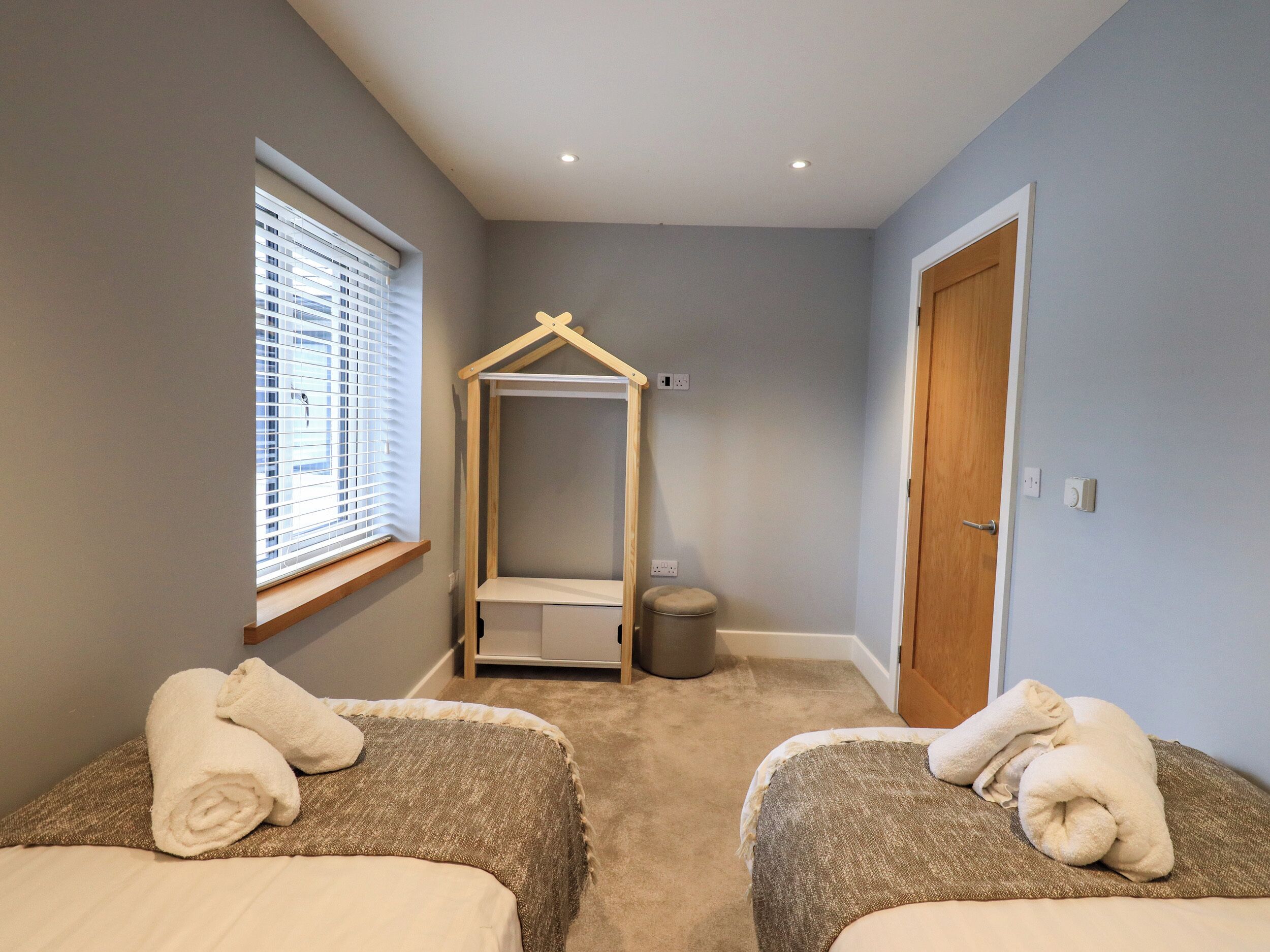A small, well-lit bedroom with two beds, a window with blinds, a wooden hanging chair, and a door.