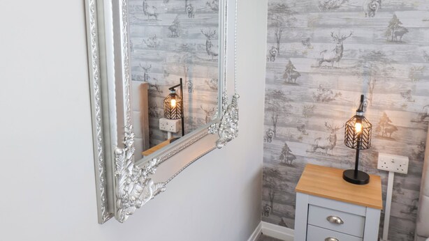 A mirror with an ornate frame, a wooden side table with a lamp, and a wall with a nature-themed wallpaper. - 315 Filey Road (Scarborough)
