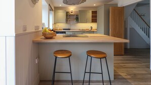 A modern kitchen with a central island, two bar stools, wooden flooring, and light-colored cabinetry. - Belfry Cottage (Colyton)