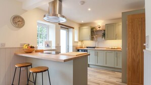 A modern kitchen with a central island, bar stools, a built-in oven, and a large window. - Belfry Cottage (Colyton)