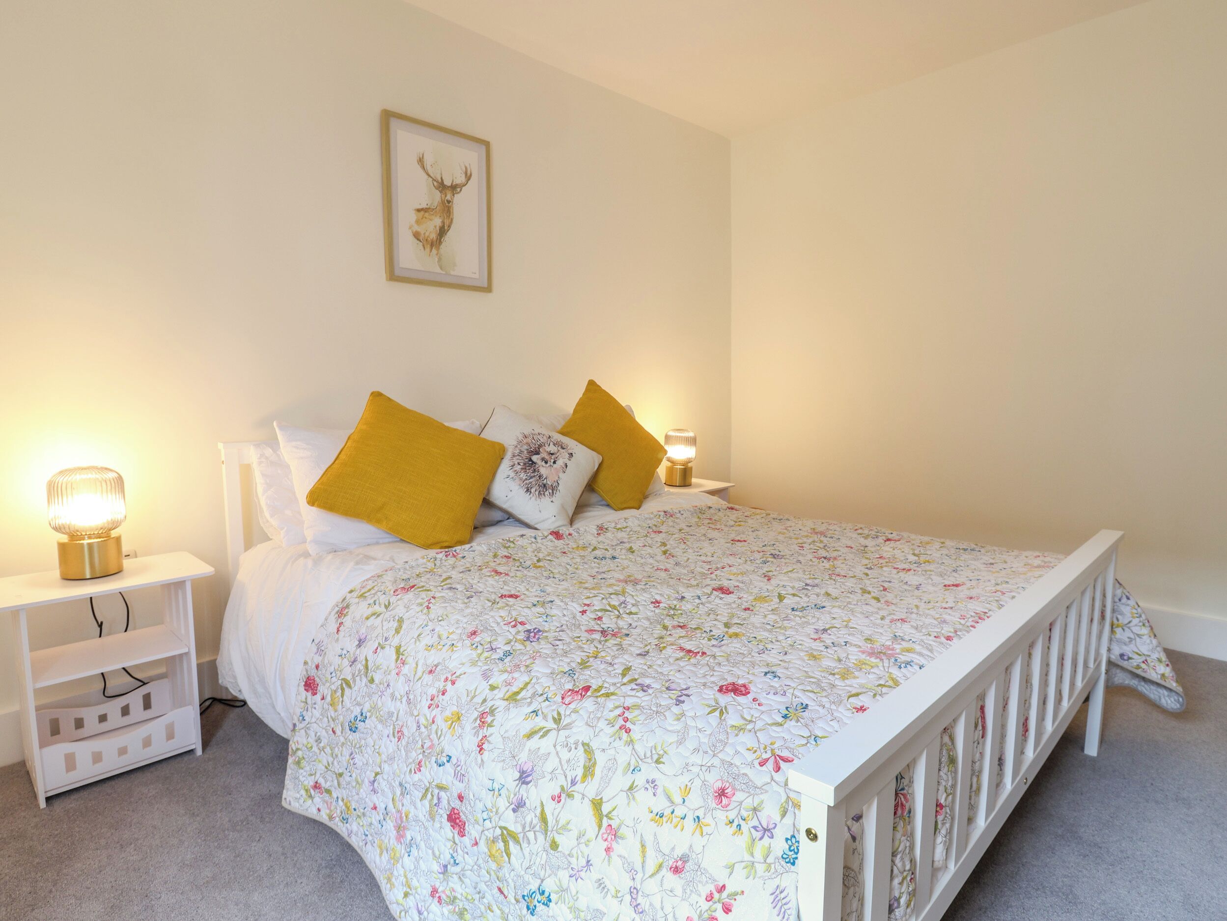 A bedroom with a floral bedspread, two bedside lamps, a framed deer artwork, and a white crib.