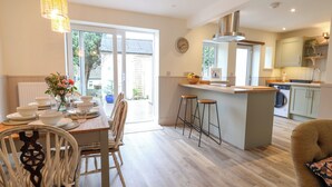 A modern kitchen with a central island, bar stools, and a dining area with a table set for four. - Belfry Cottage (Colyton)