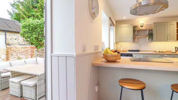 A modern kitchen with a wooden island, bar stools, and a dining area with wicker furniture. - Belfry Cottage (Colyton)