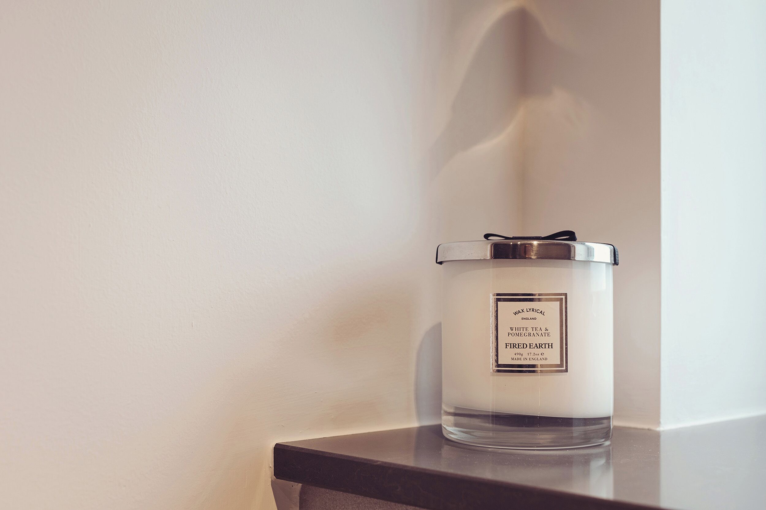 A clear glass candle with a metal lid on a dark surface against a plain wall.