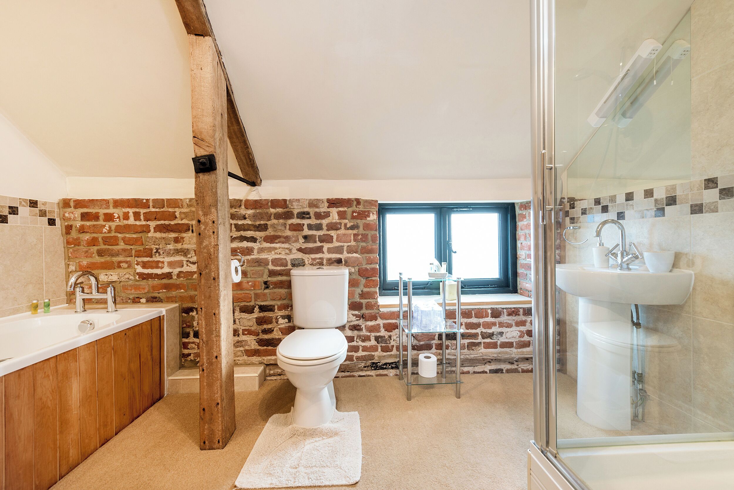 Un baño con pared de ladrillo, inodoro blanco, cabina de ducha con mampara de cristal y tocador de madera.