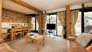 A cozy living room with a brick wall, wooden furniture, a TV, and large windows with curtains. - Snipe (Beccles)